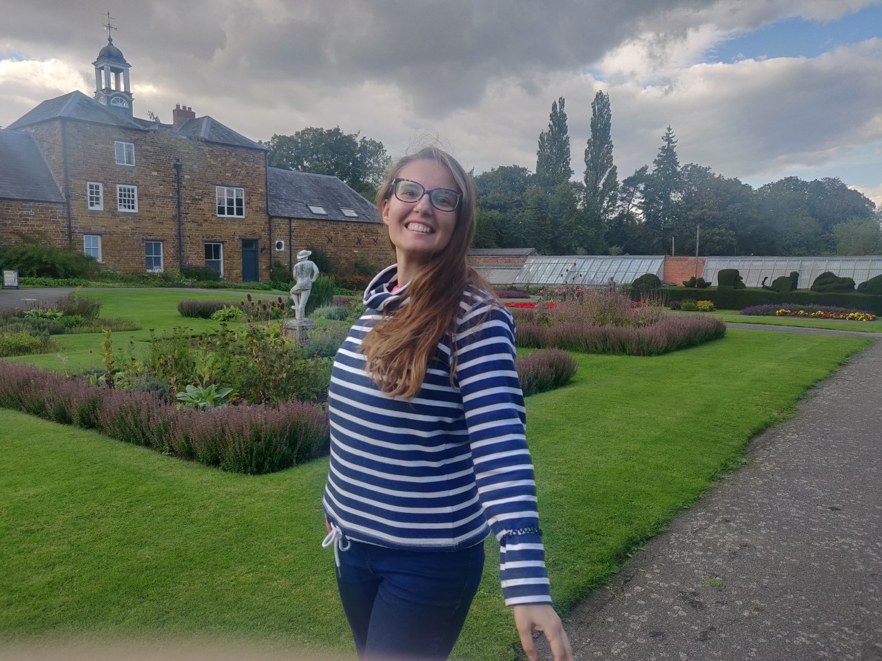 A young woman is smiling in a large garden. She is smiling at the camera.