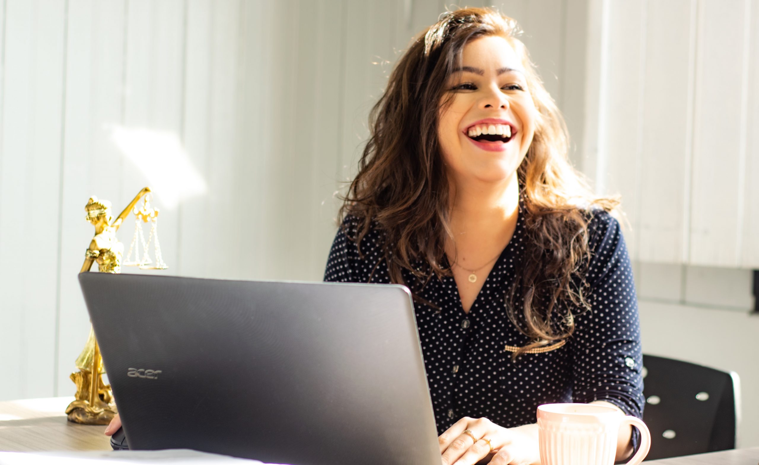 A young woman sat at a desk with a laptop in front of her. She is laughing joyfully.