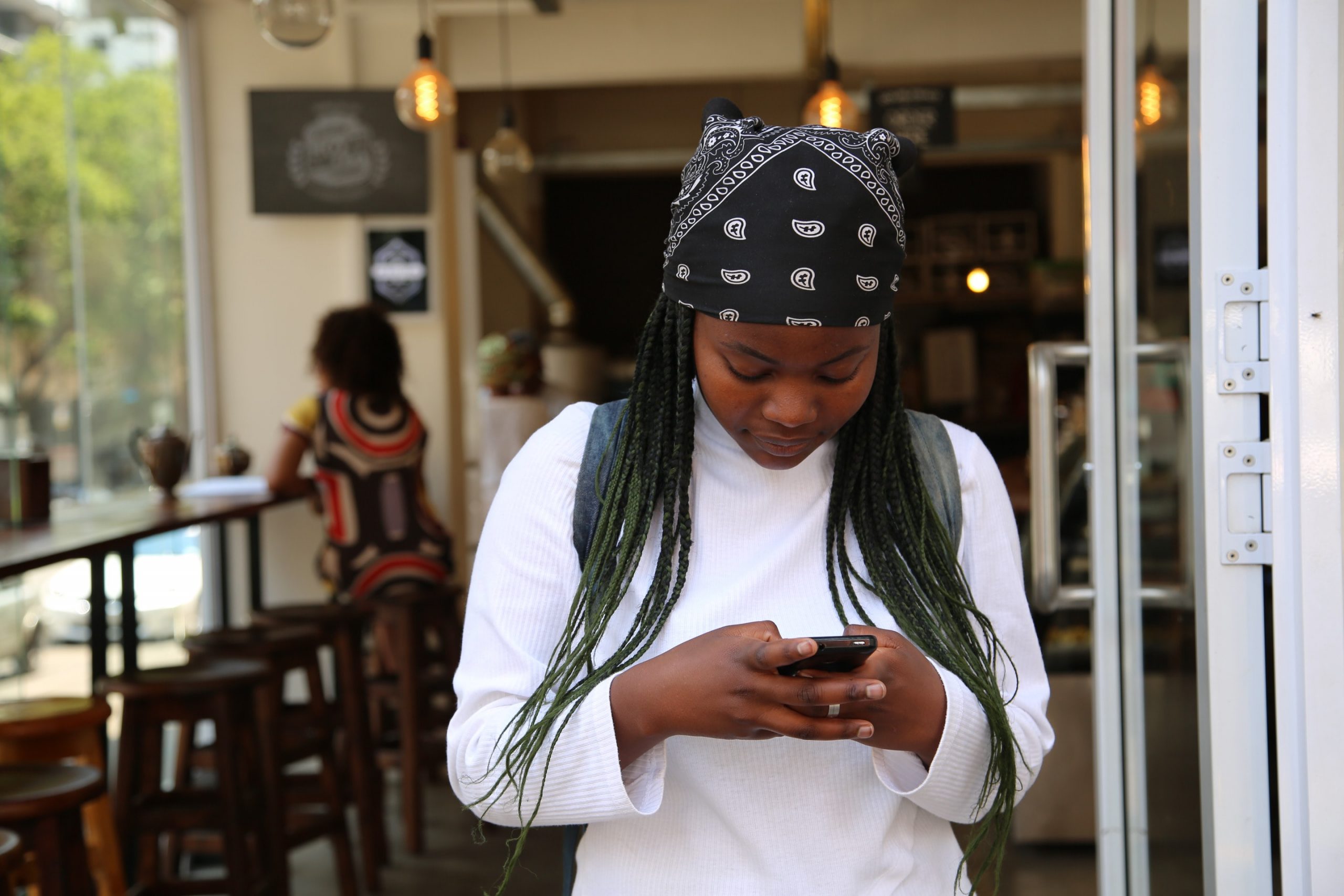 A young black woman wearing a bandana looking down at her mobile phone, texting. In the background is a restaurant or bar.