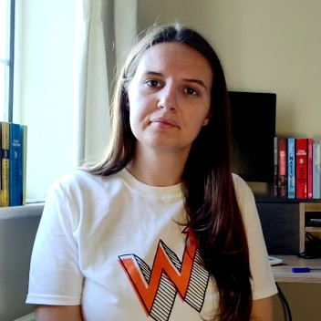 Iulia wears a 'Young Women's Trust' branded T-shirt in a photo. She is sat indoors and has a serious expression on her face.