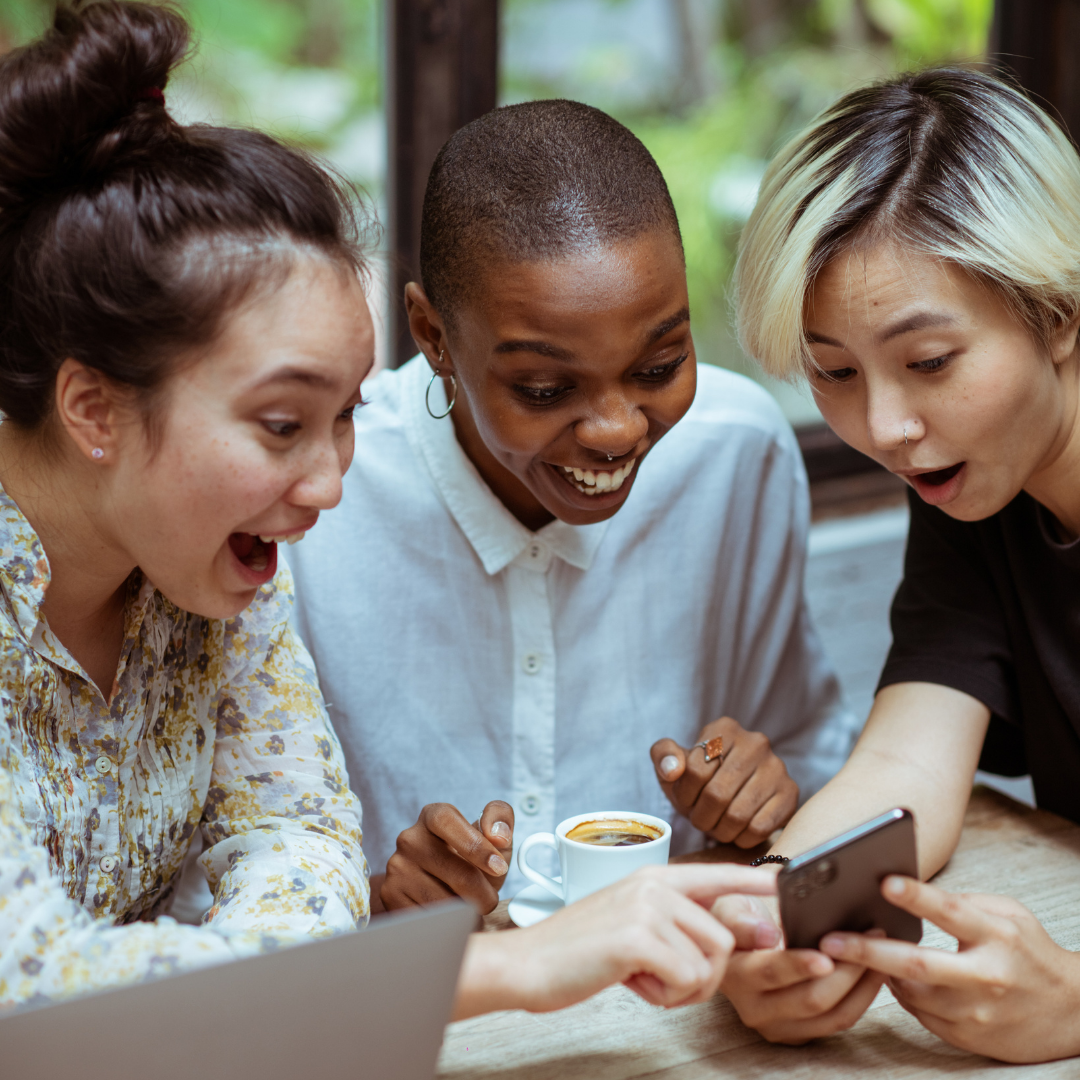 3 young women ait around a table. They are all staring excitedly at the same phone