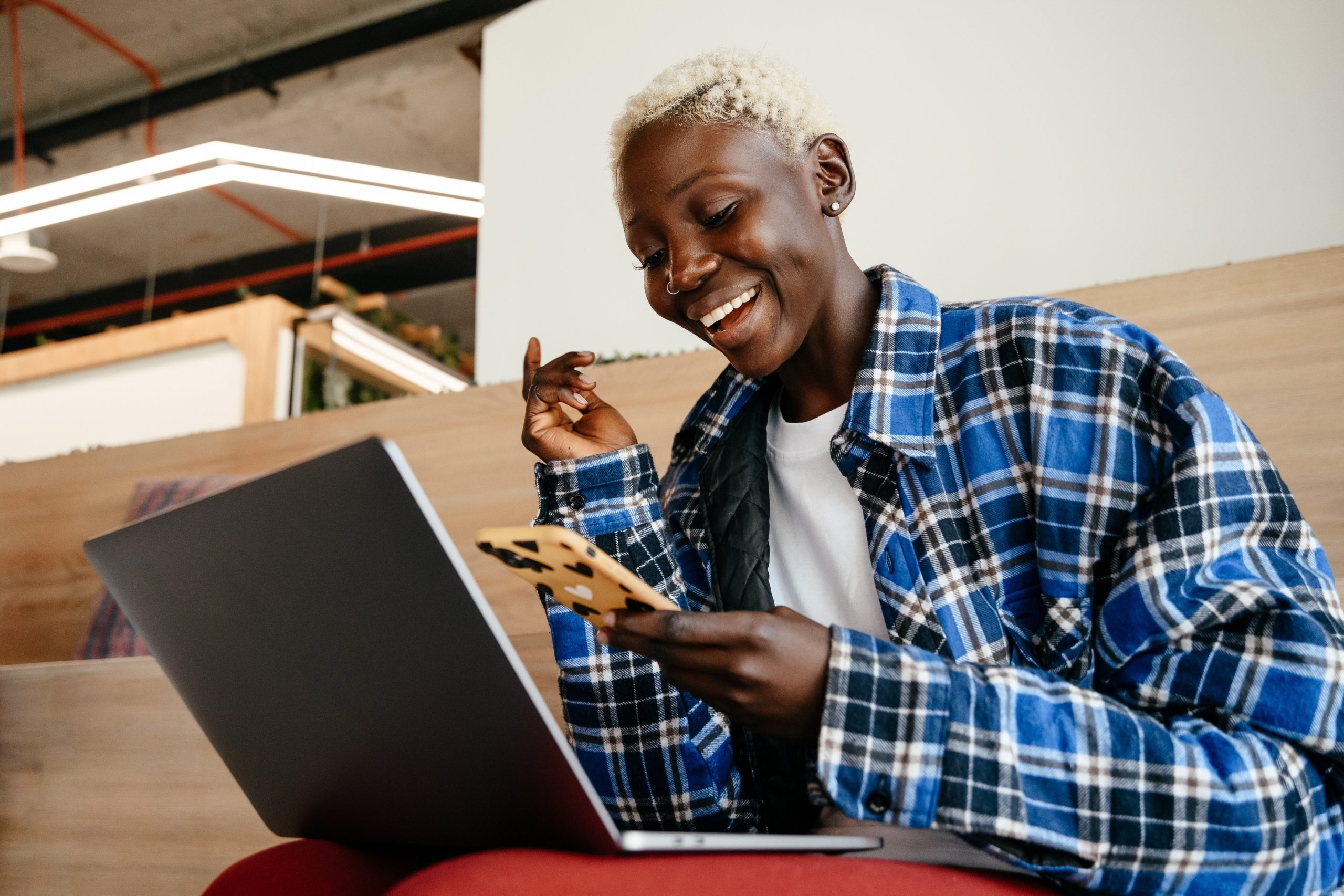 A young woman is sitting with a laptop on her lap. She is lookng at her phone which is in her hand, with a huge smile on her face.