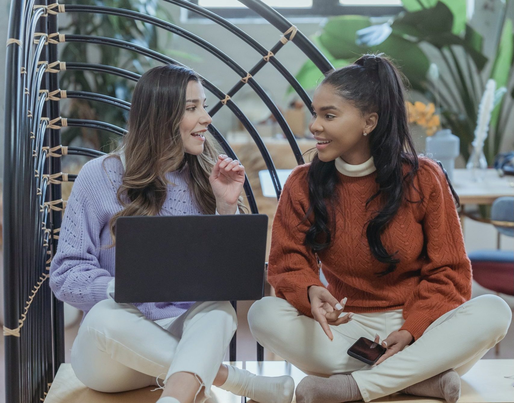 Two young women sit with a laptop in front of them. They look at each other and smile.