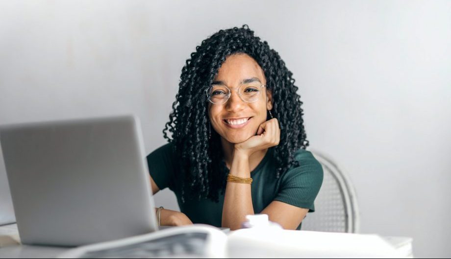 A young woman sits by her open laptop, she is looking at the camera and grinning