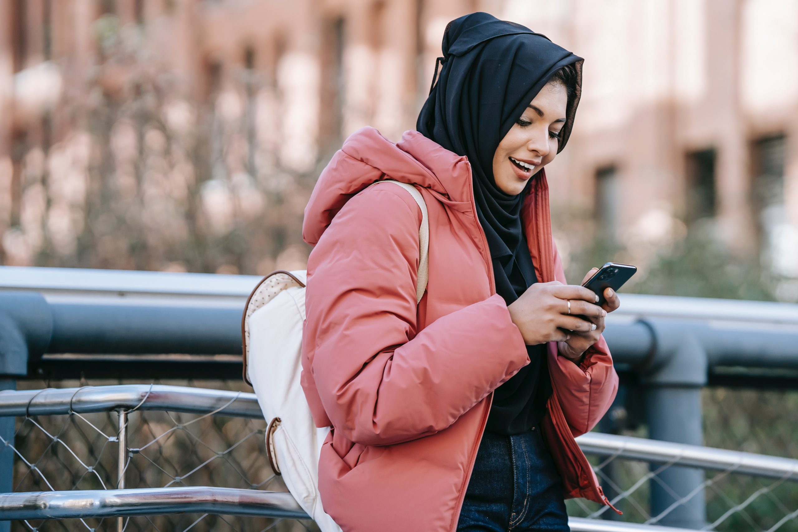 A young woman is outside, she wears a warm puffa jacket and is smiling looking down at her phone.