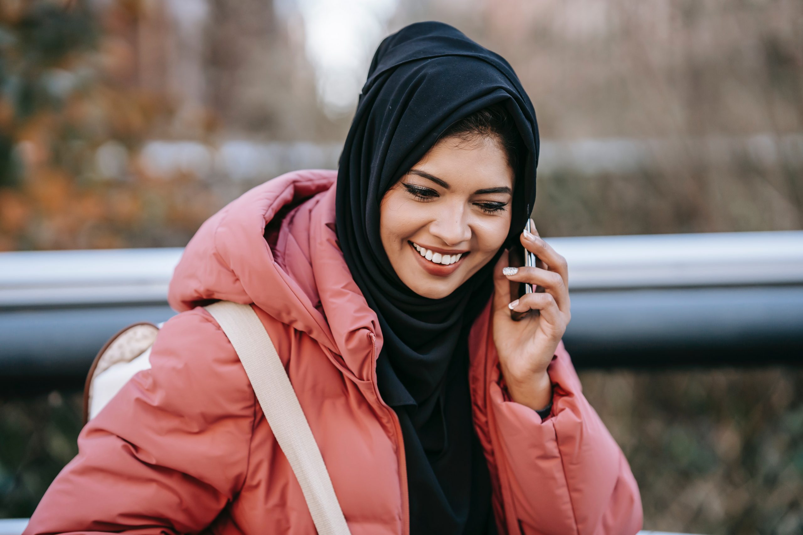 A young woman in a headscarf stands outside. She is holding a smartphone to her ear and smiling broadly.