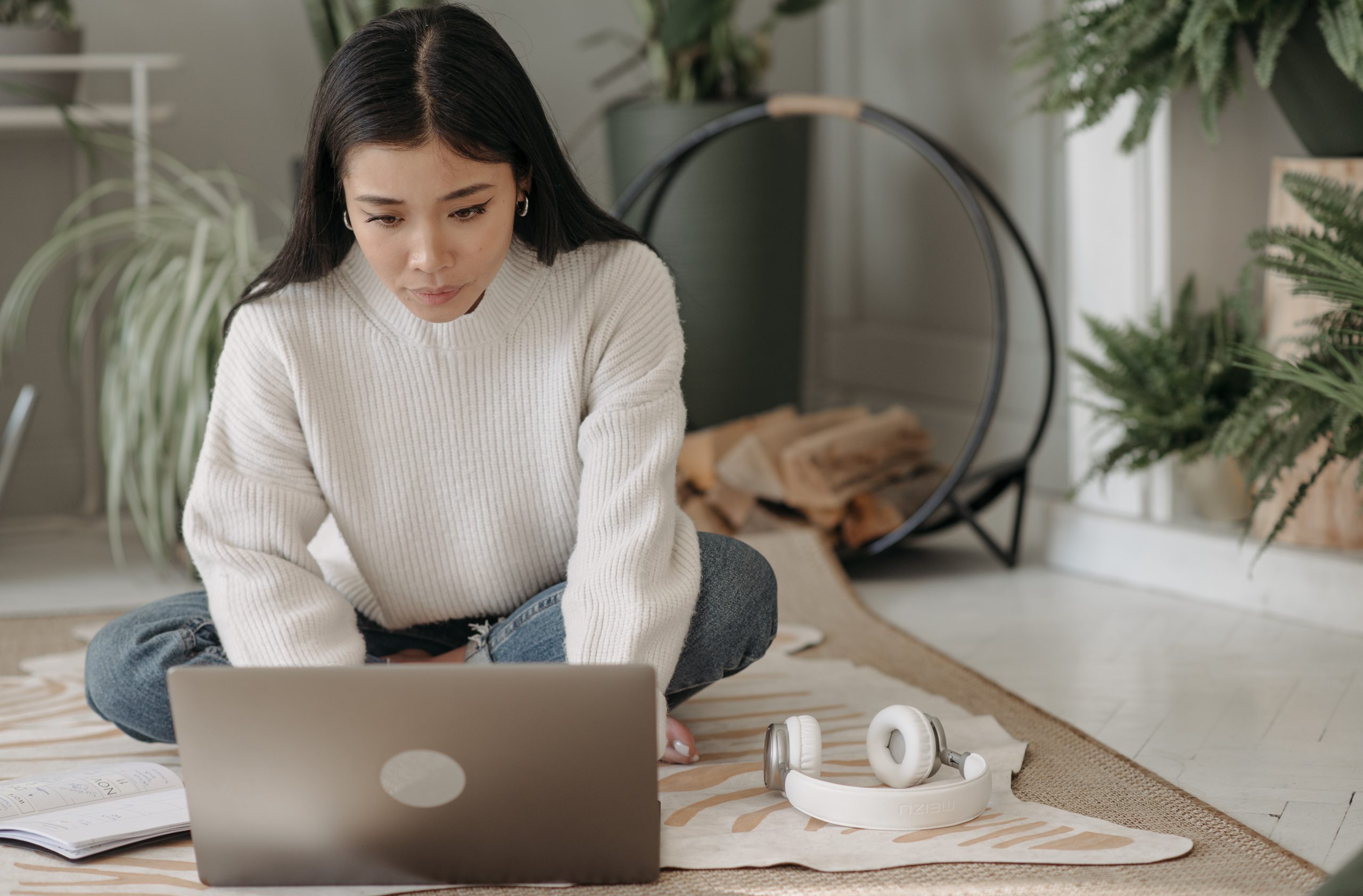 A young woman sits on the floor, she is looking down at a laptop and typing on it.