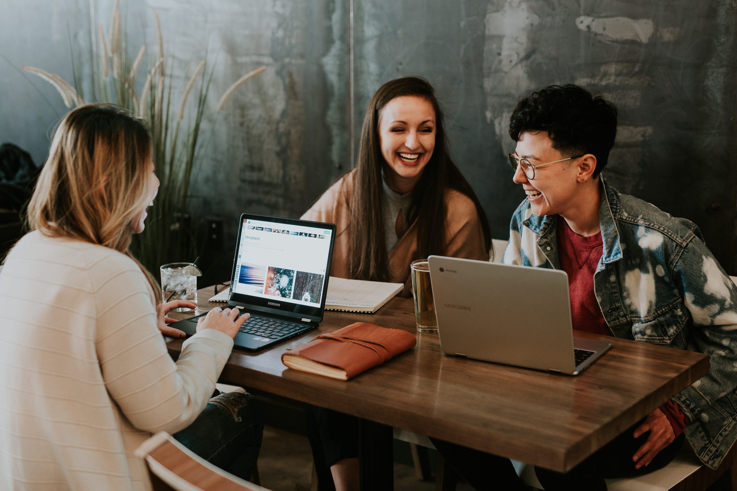 3 people are sitting at a table looking at laptops and smiling with each other