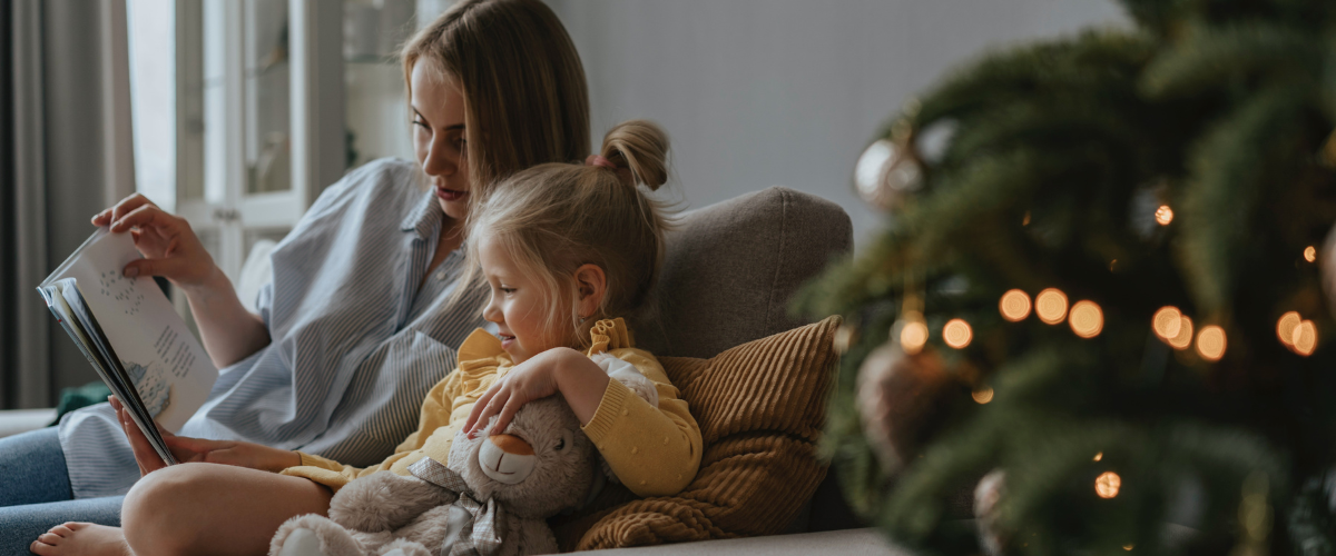 A woman and her young child are sitting on a sofa reading a book. To their right is a Christmas tree