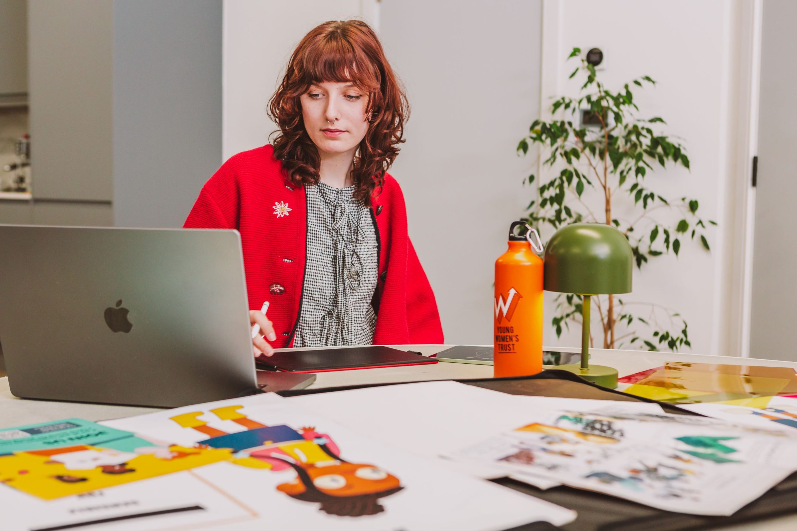 The photo shows a young woman at work, sitting on a desk and staring into a laptop