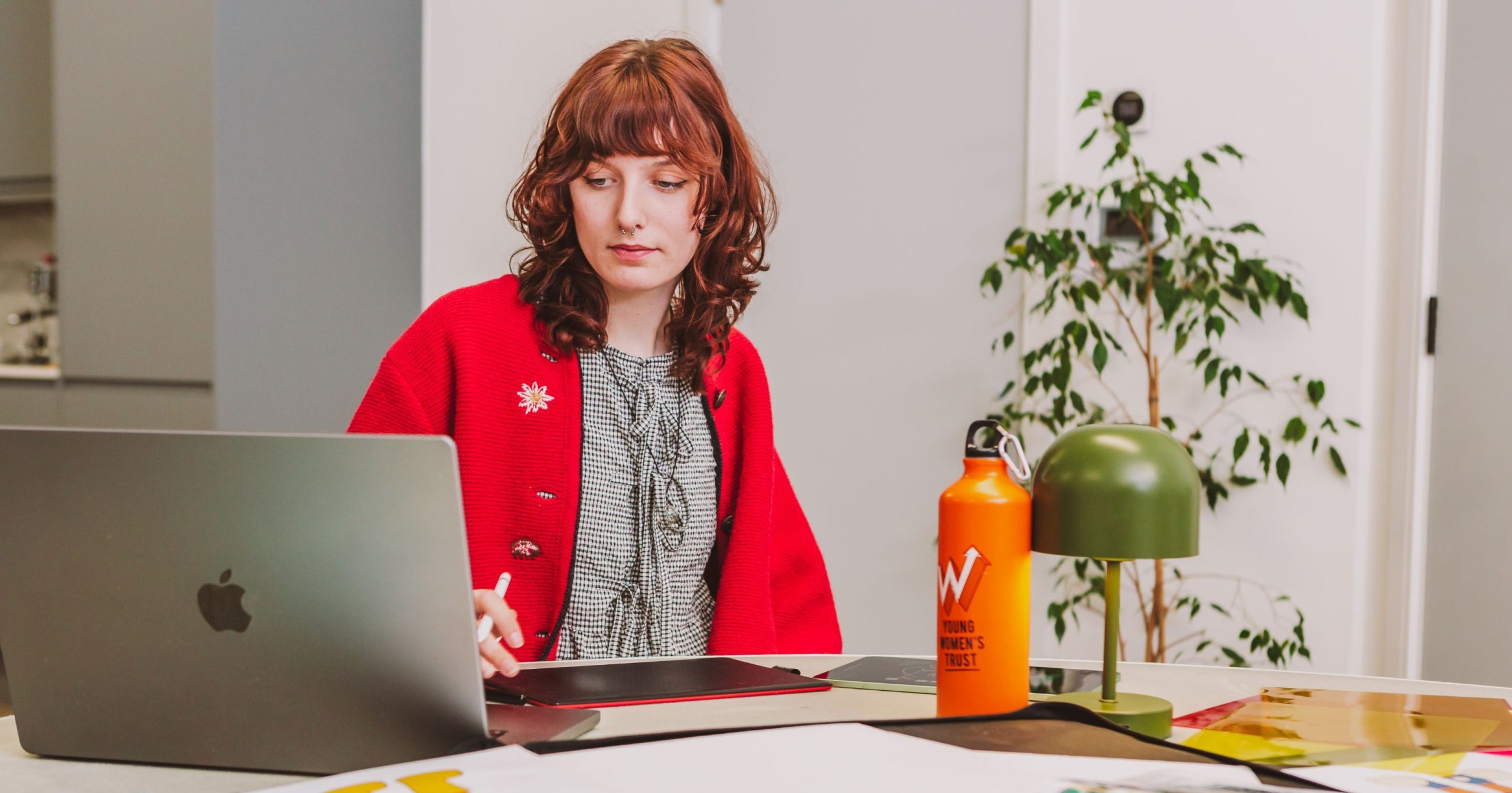 The photo shows a young woman at work, sitting on a desk and staring into a laptop