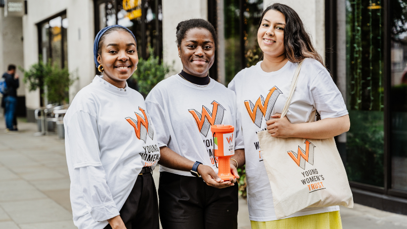three young women wearing Young Women's Trust t-shirts, holding a branded collection tin. They are smiling at the camera.