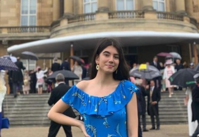 Jade stands on the steps of Buckingham Palace with one hand on her hip, whilst people file into the building behind her. She looks happy and radiates confidence.