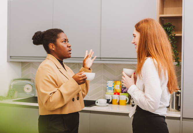 two women having a conversation in the kitchen of an office, holding cups of tea
