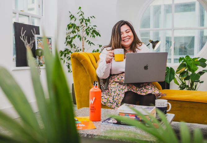 woman smiling at her laptop screen, holding a cup of tea