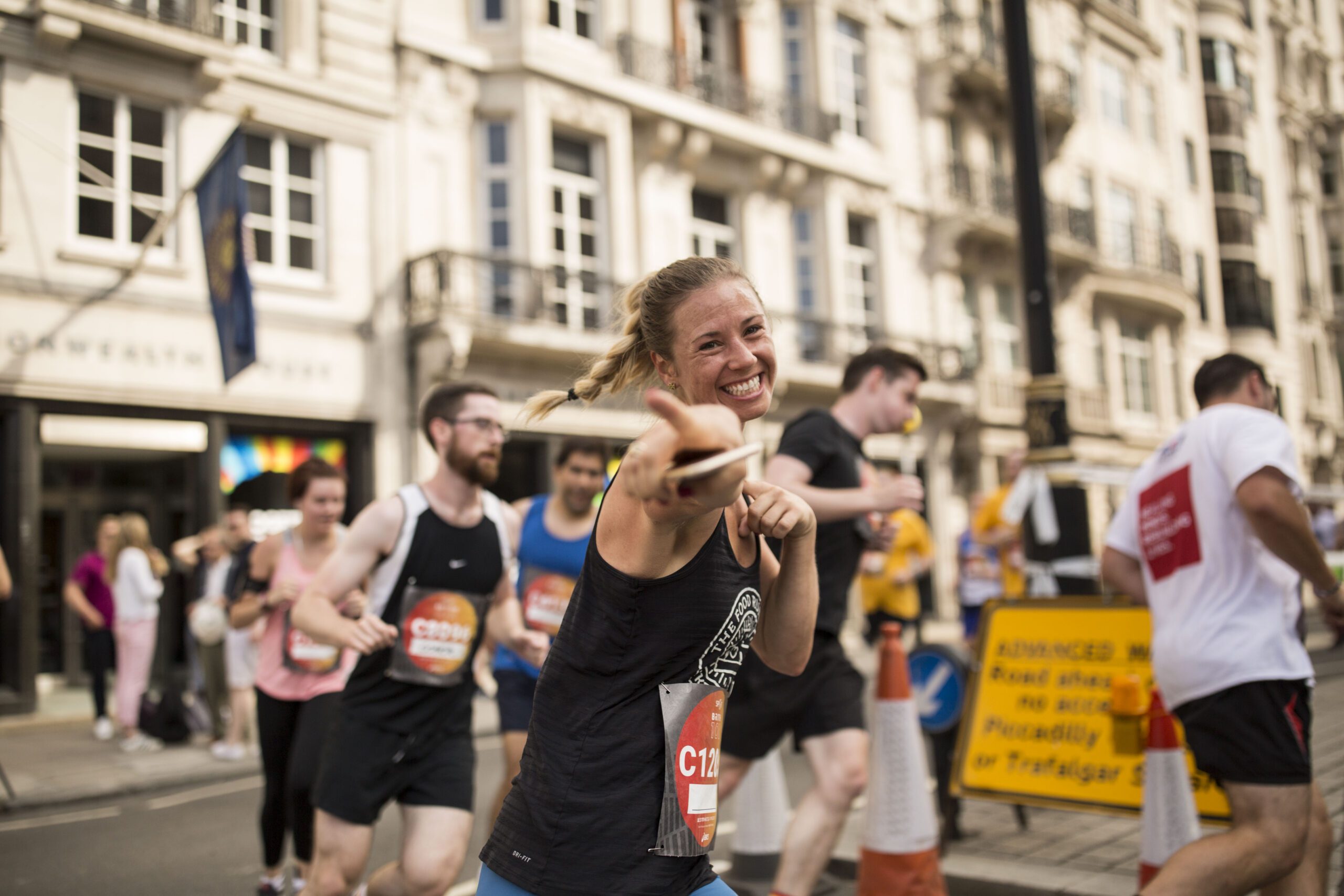 people running a marathon in blur, with camera focus on a young woman who is smiling and pointing her finger towards the camera